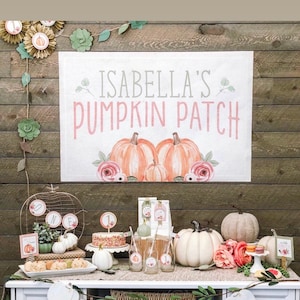 May include: A white table with a wooden top and white drawers. The table is decorated with a garland of green leaves and white pumpkins. There is a sign on the wall that says "Isabella's Pumpkin Patch". There are also several other decorations on the table, including a cake, cupcakes, and a variety of other fall-themed items.