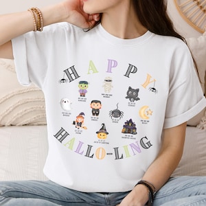 May include: White t-shirt with a colorful Halloween-themed graphic. The design features the words "HAPPY HALLO-LING" in a circular arrangement, along with various cartoon characters like a ghost, mummy, and spider.