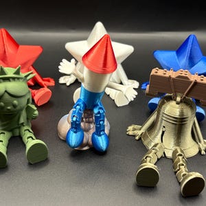 May include: A collection of five articulated figurines including a green Statue of Liberty, a red star, a white star, a blue star, and a metallic gold Liberty Bell. A central rocket-shaped figure features a red cone, white body, and blue base.