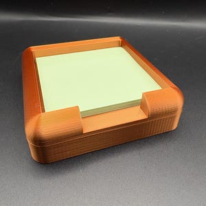 May include: A square, copper-colored plastic holder with rounded corners containing a stack of light green square sticky notes. The holder features a low-profile frame with a cutout section on the front edge to allow for easy access to the paper notes.
