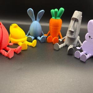 May include: A collection of colorful 3D-printed figurines, including a red tulip, yellow chick, blue bunny, orange carrot, gray Moai statue, and purple bunny. Each figure has articulated legs and arms, sitting on a dark surface.