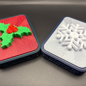 May include: Two decorative boxes with festive designs. One features a green holly and red berries on a red background. The other has a white snowflake on a silver background. Both have dark blue borders.