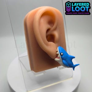 May include: A novelty earring featuring a blue and white cartoon shark biting a human ear. The ear is a flesh-toned prosthetic. The image includes the text "LAYERED LOOT" and "CURATED CHAOS. PRINTED CHARM."