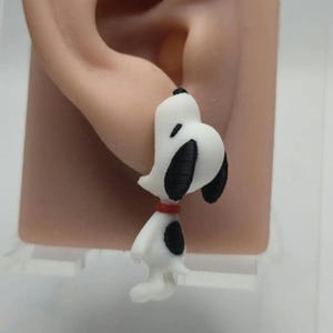 May include: A white and black Snoopy earring with a red collar. The earring is a small, three-dimensional figure of the cartoon character. The earring is attached to an ear.