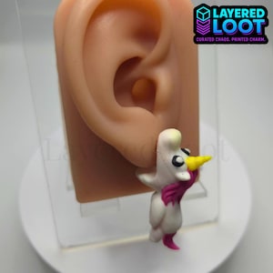 May include: A whimsical white and pink duck earring with a yellow beak and black eyes. The earring is attached to a prosthetic ear. The text "LAYERED LOOT" is visible in the upper right corner.