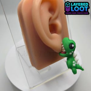 May include: A green dinosaur earring with white accents is shown on a display ear. The earring has a cartoonish design with a wide open mouth. The logo "LAYERED LOOT" is in the upper right corner.