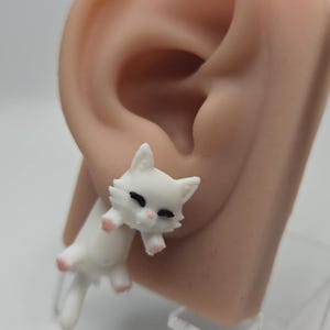 May include: A white cat-shaped earring with black eyes and pink paws. The earring is attached to a flesh-toned ear. The cat is in a playful pose, with its tail and paws extended. The earring is made of a smooth, glossy material.