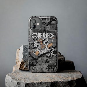 Könnte beinhalten: Schwarz-weißes Handycase mit Manga-Design. Es zeigt den Schriftzug "ONE PIECE" und eine Charakterillustration. Das Case hat eine glänzende Oberfläche und wird auf einer Steinoberfläche präsentiert.