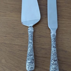 Könnte beinhalten: Ein silbernes Tortenheber- und Messerset. Die Klingen sind schlicht, während die Griffe ein verziertes Blumenmuster aufweisen. Das Set wird auf einer Holzoberfläche präsentiert.