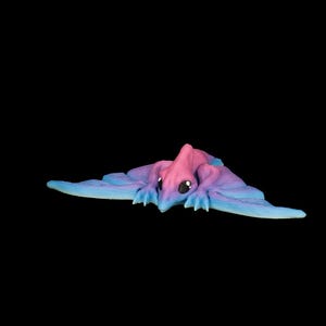 Puede incluir: Una pequeña figura de criatura articulada que se asemeja a una mantarraya con un esquema de color degradado que transiciona de rosa en la cabeza a morado y azul a lo largo de las alas anchas y planas. La figura tiene ojos grandes, oscuros y brillantes.