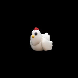 Può includere: Una statuetta di pollo bianco stampata in 3D con cresta rossa, occhi neri e becco giallo. Il pollo è seduto con le ali leggermente aperte. Lo sfondo è nero.