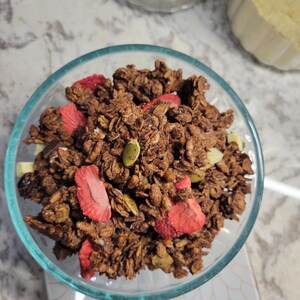 May include: A clear glass bowl filled with chocolate granola. The granola contains dried strawberries, pumpkin seeds, and white chocolate chunks. The granola is a mix of brown and tan colors.