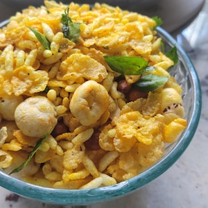 May include: A clear glass bowl filled with a savory snack mix containing yellow corn flakes, puffed rice, roasted peanuts, and round puffed lotus seeds, all seasoned with a vibrant yellow spice blend and garnished with fresh green curry leaves.