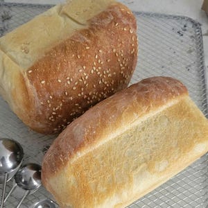 May include: Two rectangular loaves of baked bread resting on a wire cooling rack. One loaf features a golden-brown crust topped with scattered sesame seeds, while the other loaf has a smooth, lighter-colored crust. Three silver measuring spoons.