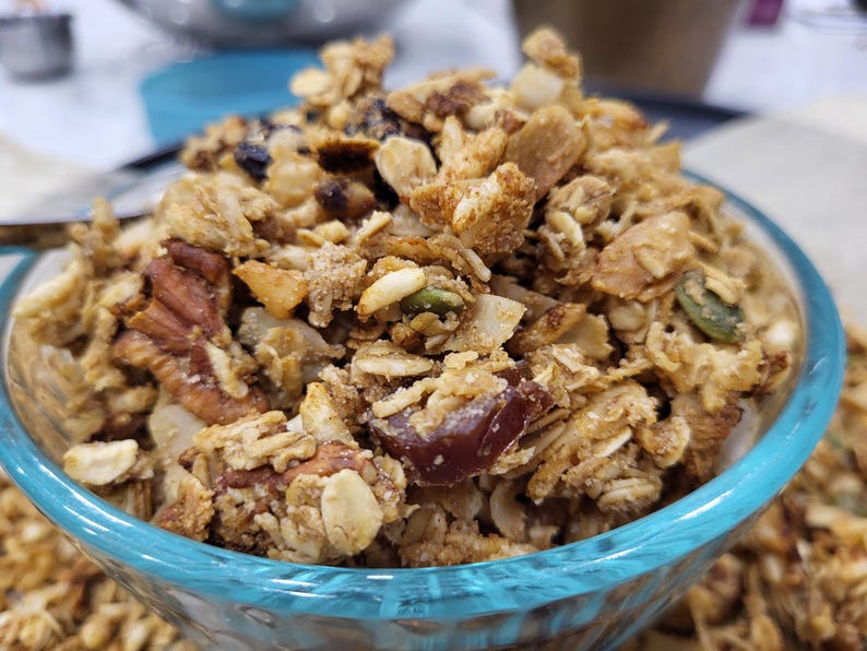 May include: A close-up of a clear glass bowl filled with homemade granola. The granola is a mix of golden-brown oats, nuts, seeds, and dried fruit. The bowl has a light blue rim and sits on a white surface.