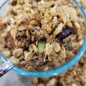 May include: Close-up of a spoonful of granola with oats, nuts, seeds, and dried fruit. The granola is a mix of golden brown and tan colors, with visible pieces of pumpkin seeds and dried cranberries. A clear glass bowl with a blue rim is in the background.
