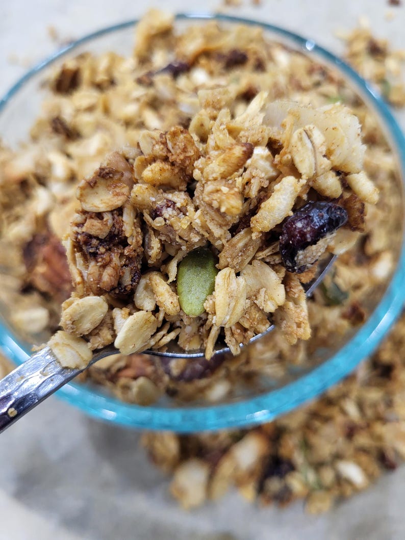 May include: Close-up of a spoonful of granola with oats, nuts, seeds, and dried fruit. The granola is a mix of golden brown and tan colors, with visible pieces of pumpkin seeds and dried cranberries. A clear glass bowl with a blue rim is in the background.