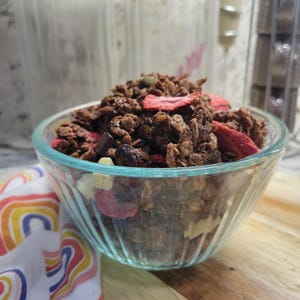 May include: A clear glass bowl filled with homemade granola. The granola is a mix of brown clusters, dried strawberries, and other ingredients. A colorful rainbow-patterned cloth is visible in the lower left corner. The bowl sits on a wooden cutting board.