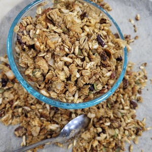 May include: A clear glass bowl filled with granola, with some granola scattered around it. The granola is a mix of golden brown and tan clusters with visible nuts and seeds. A silver spoon rests on the surface near the bowl.