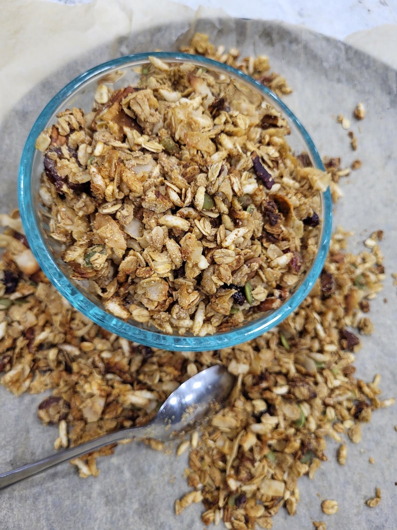 May include: A clear glass bowl filled with granola, with some granola scattered around it. The granola is a mix of golden brown and tan clusters with visible nuts and seeds. A silver spoon rests on the surface near the bowl.