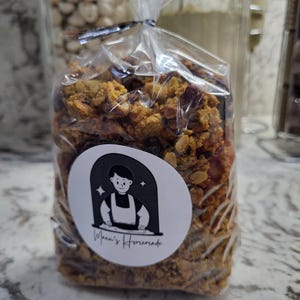 May include: A clear plastic bag filled with granola, sealed with a black twist tie. A white circular sticker with a cartoon chef and the text "Mona's Homemade" is affixed to the bag. The granola appears to be a mix of grains and dried fruit.