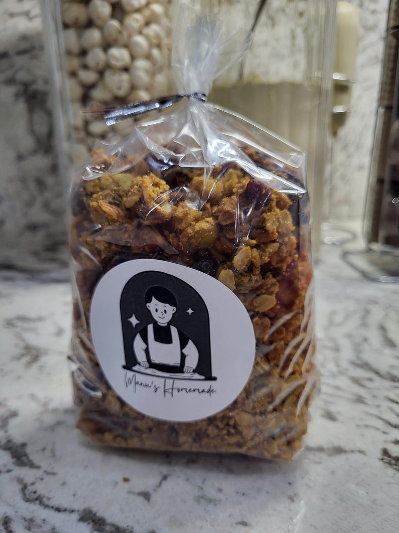 May include: A clear plastic bag filled with granola, sealed with a black twist tie. A white circular sticker with a cartoon chef and the text "Mona's Homemade" is affixed to the bag. The granola appears to be a mix of grains and dried fruit.
