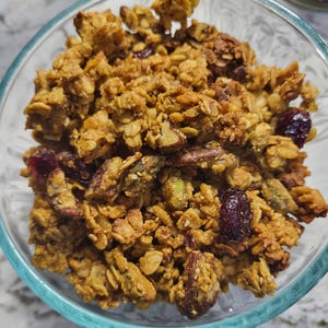 May include: A clear glass bowl filled with homemade granola. The granola is a mix of golden-brown oats, nuts, and dried cranberries. The bowl sits on a light-colored surface, with a hint of a pumpkin-themed candle in the background.