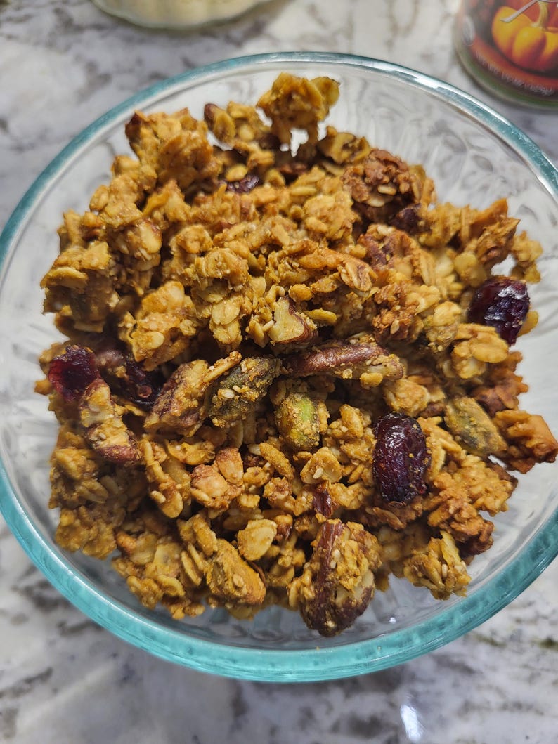 May include: A clear glass bowl filled with homemade granola. The granola is a mix of golden-brown oats, nuts, and dried cranberries. The bowl sits on a light-colored surface, with a hint of a pumpkin-themed candle in the background.