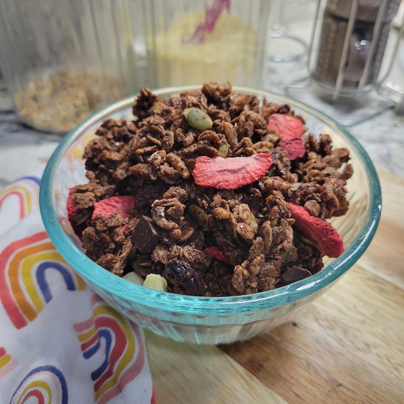 May include: A clear glass bowl filled with chocolate granola, dried strawberries, and pumpkin seeds. The granola is on a wooden surface with a rainbow-patterned cloth in the background. Other ingredients are visible in the background.
