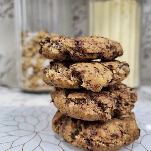 May include: A stack of four chocolate chip cookies. The cookies are golden brown with visible chocolate chips. They are stacked on a white surface with a floral pattern. Jars of ingredients are in the background.