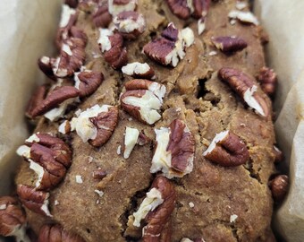 May include: A loaf of freshly baked bread, topped with pecan halves. The bread is a warm brown colour, baked in a metal loaf tin lined with parchment paper. The pecans are a rich, dark brown.