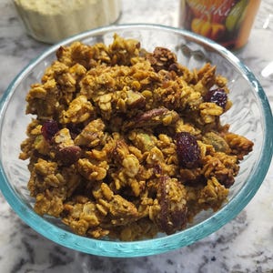 May include: A clear glass bowl filled with homemade granola. The granola is a mix of golden-brown oats, nuts, and dried cranberries. A jar of white powder and a can of "Organic Pumpkin" are in the background.