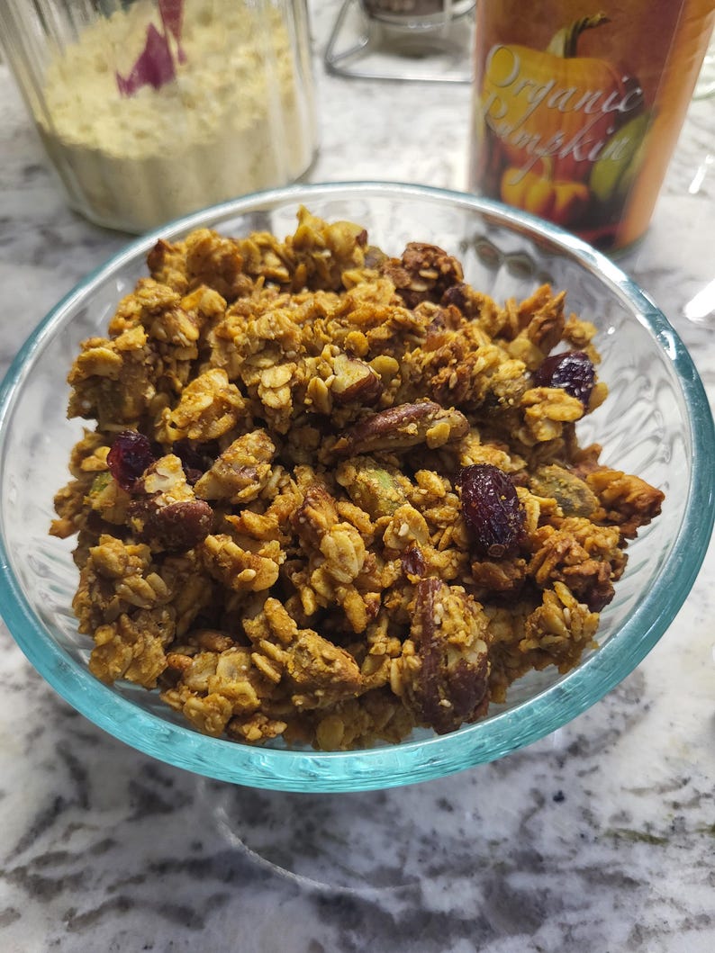 May include: A clear glass bowl filled with homemade granola. The granola is a mix of golden-brown oats, nuts, and dried cranberries. A jar of white powder and a can of "Organic Pumpkin" are in the background.