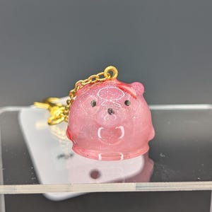 May include: A translucent pink resin keychain shaped like a small bear head with embedded glitter particles. The bear features two small black eyes, a black nose, and a gold-toned metal chain attached to the top. The surface is smooth and glossy.