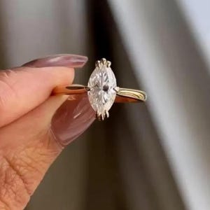 Könnte beinhalten: Ein goldener Ring mit einem Diamanten im Marquiseschliff. Der Diamant ist mit Krappen gefasst und hat ein klares, brillantes Aussehen. Das Band ist aus schlichtem, poliertem Gold. Der Ring wird hochgehalten, um das Design und den Glanz zu zeigen.