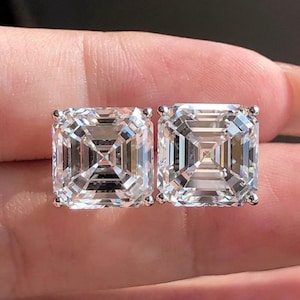 May include: Two square-cut, clear gemstone stud earrings. The gemstones are held in place by small, silver-colored prongs. The earrings are displayed against a blurred background.