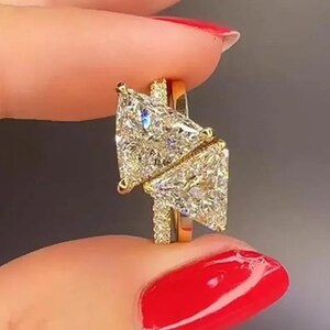 May include: A gold ring with two large, sparkling, triangular-cut diamonds. The diamonds are set in a gold band with a delicate, textured design. The ring is held up by a person's fingers with red nail polish.