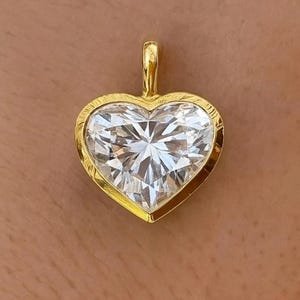Könnte beinhalten: Herzförmiger Anhänger mit einem großen, klaren Edelstein in einem goldenen Rahmen. Der Anhänger hat eine goldene Öse zum Anbringen an einer Kette. Der Edelstein ist facettiert und erzeugt einen funkelnden Effekt. Schmuckstück.