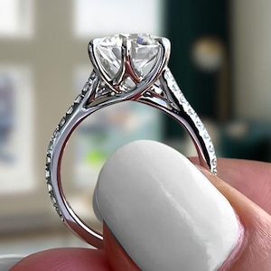 May include: A silver engagement ring featuring a large, clear, round-cut diamond. The band is set with smaller diamonds. The ring is held up against a blurred background.