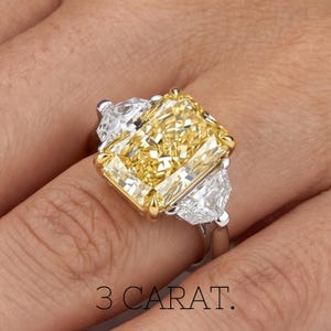 May include: A radiant, yellow diamond ring with a rectangular center stone, flanked by two white, triangular diamonds. The ring has a gold setting around the center stone and a silver band. The text "3 CARAT" is at the bottom.