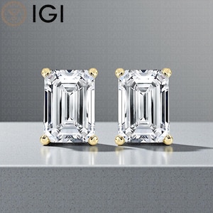 May include: A pair of emerald-cut diamond stud earrings with gold settings. The earrings are displayed on a grey surface. The IGI logo is visible in the upper left corner.