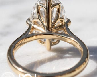 May include: A gold ring featuring a large oval-cut clear gemstone set in a decorative, leaf-inspired metal basket setting. The band is smooth and polished, showcasing a high-shine finish that reflects light across the gold surface and the stone.