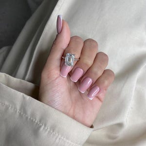 May include: A silver ring with a large, rectangular-cut diamond. The ring is displayed on a hand with pink nails and white tips. The background is a neutral color.