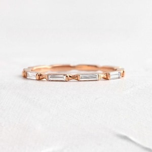 May include: A rose gold ring with a series of rectangular, clear gemstones set around the band. The ring has a delicate design, with each gemstone separated by a small, decorative element. The ring is shown on a white surface.