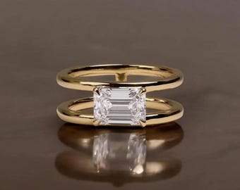 May include: A gold ring with a rectangular, clear gemstone set between two parallel bands. The ring is displayed on a reflective surface, showcasing its design and the gemstone's facets. The ring's design is modern and elegant.