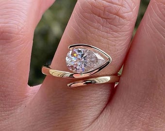 May include: A gold ring with a pear-shaped, clear gemstone. The ring's band wraps around the finger in a spiral design. The gemstone is set in a bezel setting, and the ring is shown on a finger.