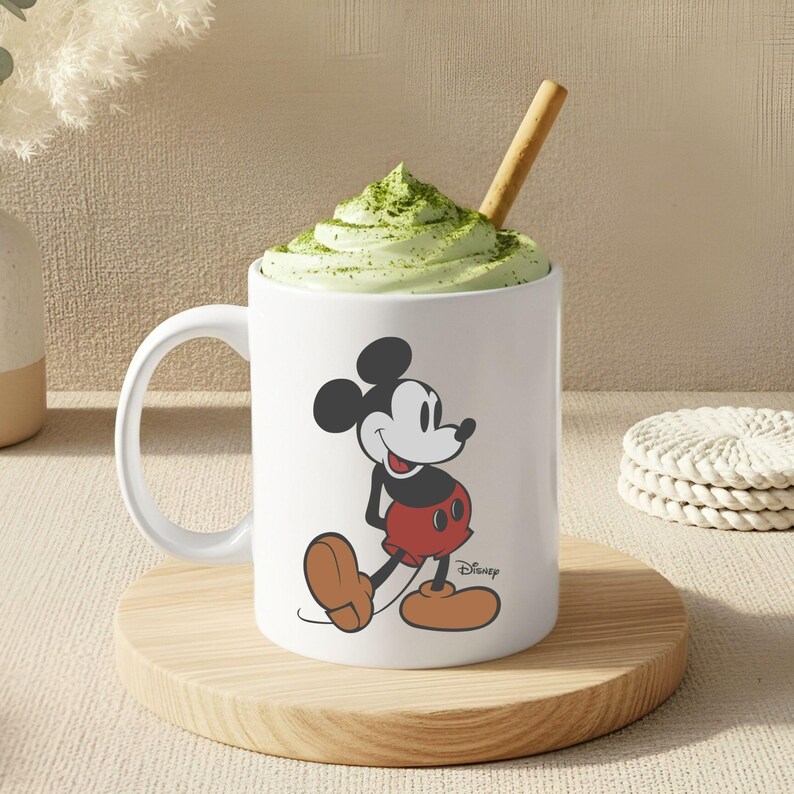 May include: A white ceramic mug featuring a classic illustration of Mickey Mouse with black ears, a red pair of shorts, and tan shoes. The mug is filled with a light green whipped topping sprinkled with green powder and includes a wooden stirring stick.