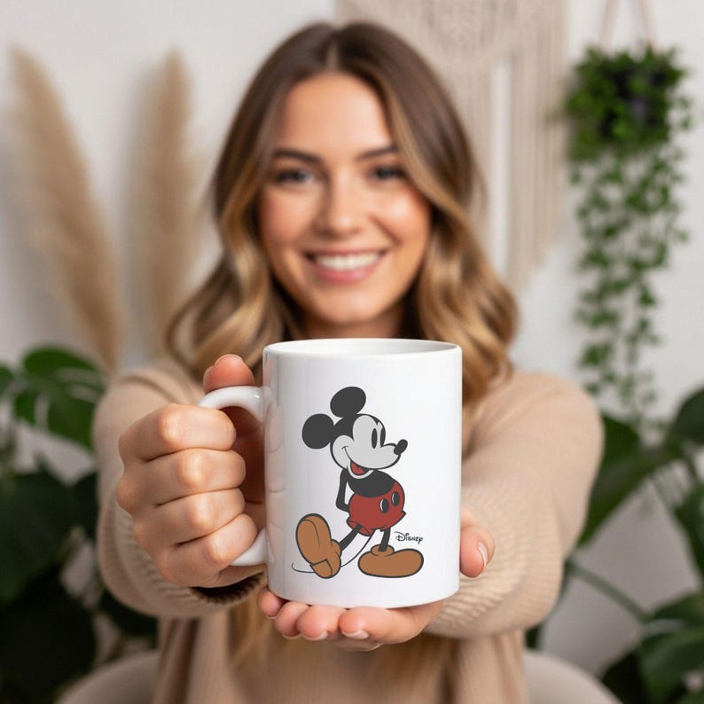 May include: A white ceramic mug featuring a classic illustration of Mickey Mouse. The character is depicted in a vintage style with a black head, red shorts, and tan shoes. The word Disney is printed in a small black script font near the character's feet.
