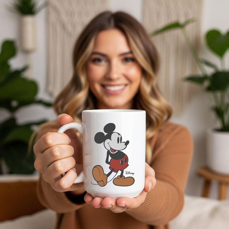 May include: A white ceramic mug featuring a classic illustration of Mickey Mouse. The character is depicted in a standing pose with black ears, a grey face, red shorts, and large brown shoes. The word Disney is printed in small black script.