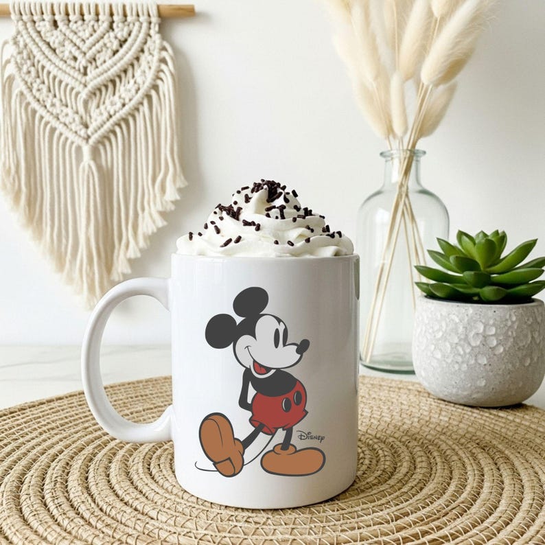 May include: A white ceramic mug featuring a classic illustration of Mickey Mouse with black ears, a red pair of shorts, and tan shoes. The mug is filled with whipped cream topped with dark chocolate sprinkles and includes the Disney logo text.